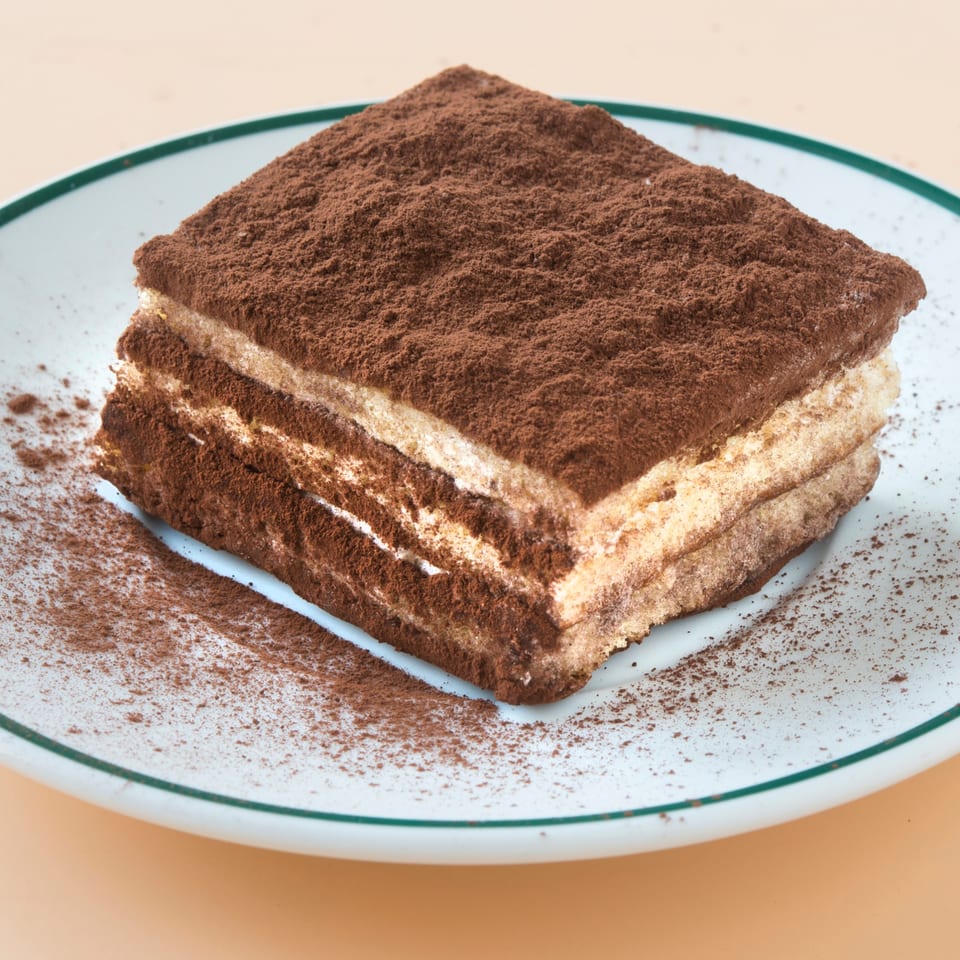 A square slice of tiramisu with layers of cream and cake, topped with cocoa powder.